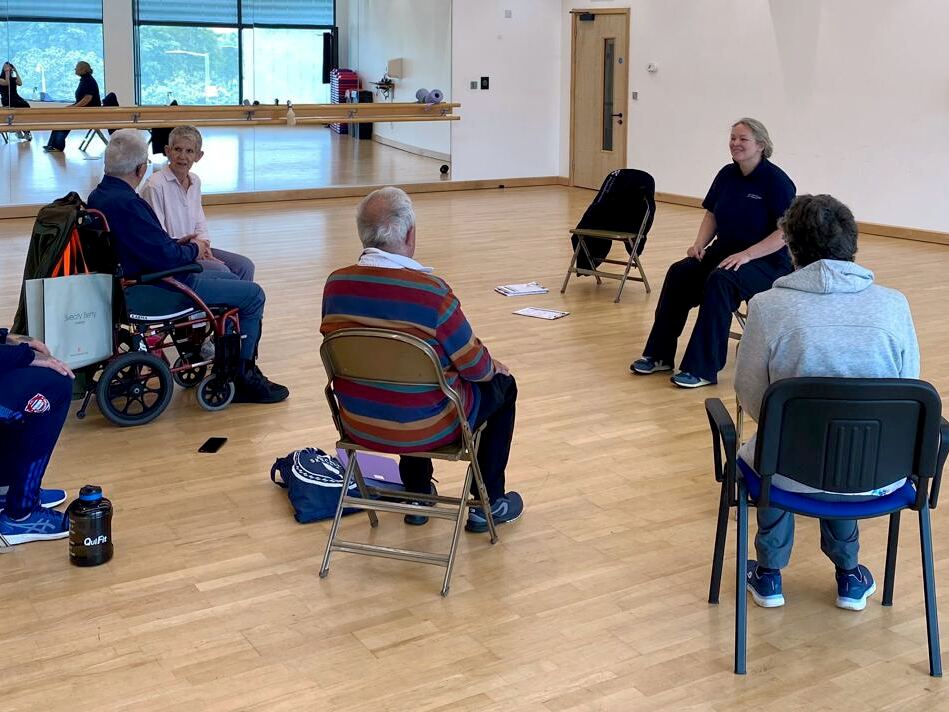 A neurological rehabilitation class: participants seated in a circle in a community hall, led by a physiotherapist.